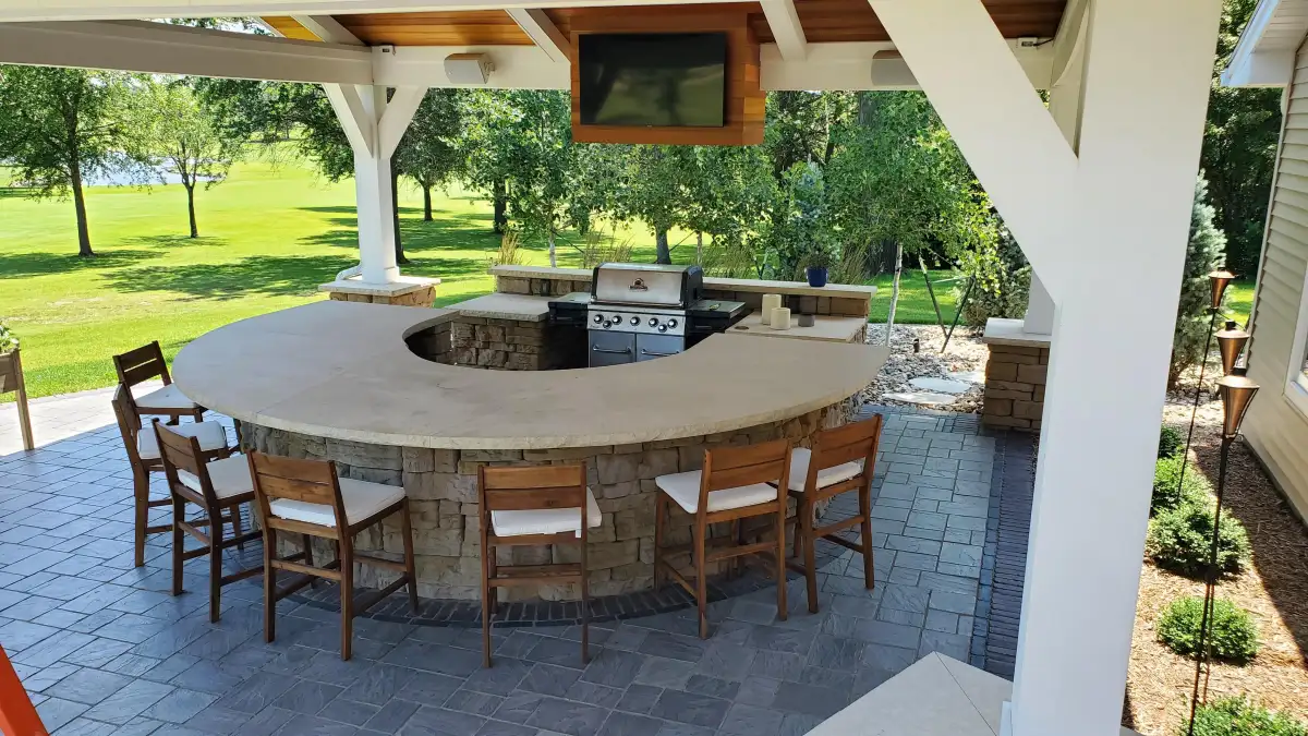 Covered outdoor kitchen with curved stone bar island and built-in grill by Matthias Landscaping